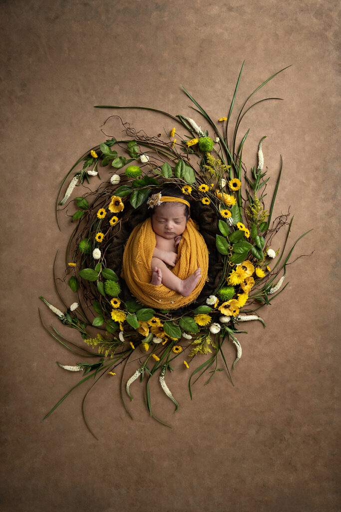 Newborn girl wrapped in mustard yellow laying int twig nest and yellow flowers