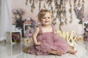 12 month old girl smiles in pretty pink dress at her floral themed cake smash