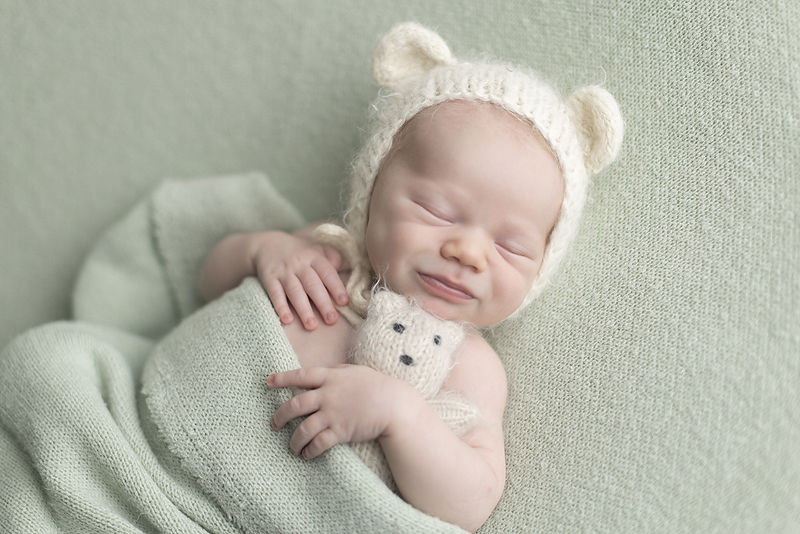 Newborn boy smiles snuggling white teddy bear while laying on green fabric