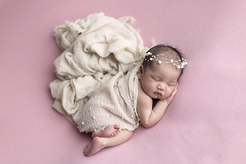 Newborn girl poses on pink fabric with cream colored scarves flowing behind her