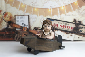 6 month old boy poses in wooden airplane against vintage aviation backdrop
