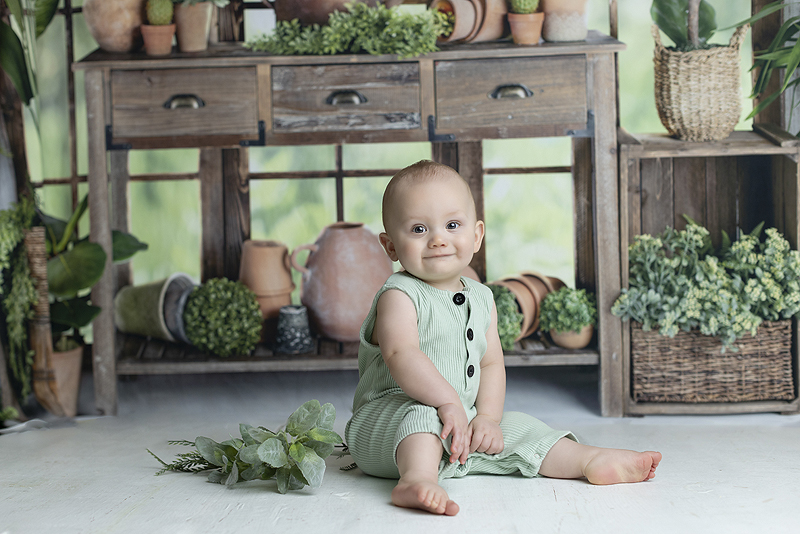 7 month old boy smiles for camera surrounded by greenery in green house